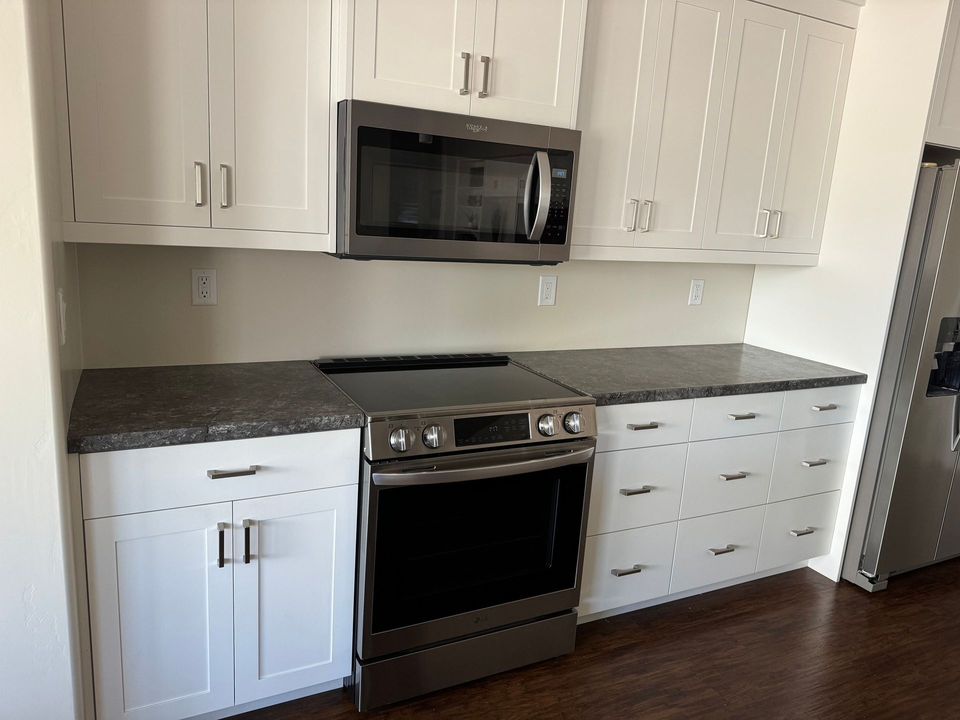 A modern kitchen featuring white cabinets, dark gray granite countertops, stainless steel appliances, and wood flooring.