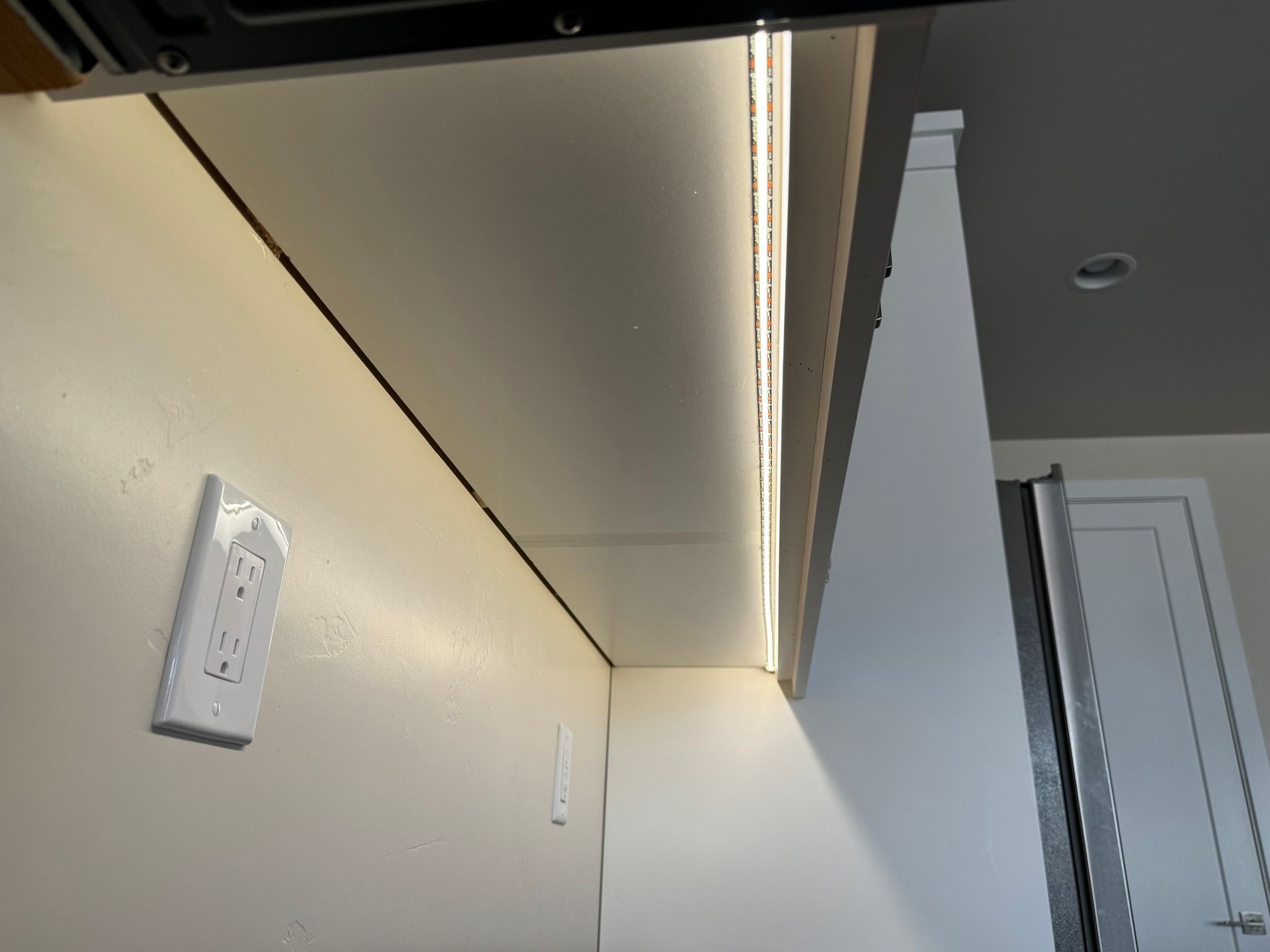 Low-angle view of a kitchen cabinet underside with a warm white LED strip light, mounted against a plain cream wall.