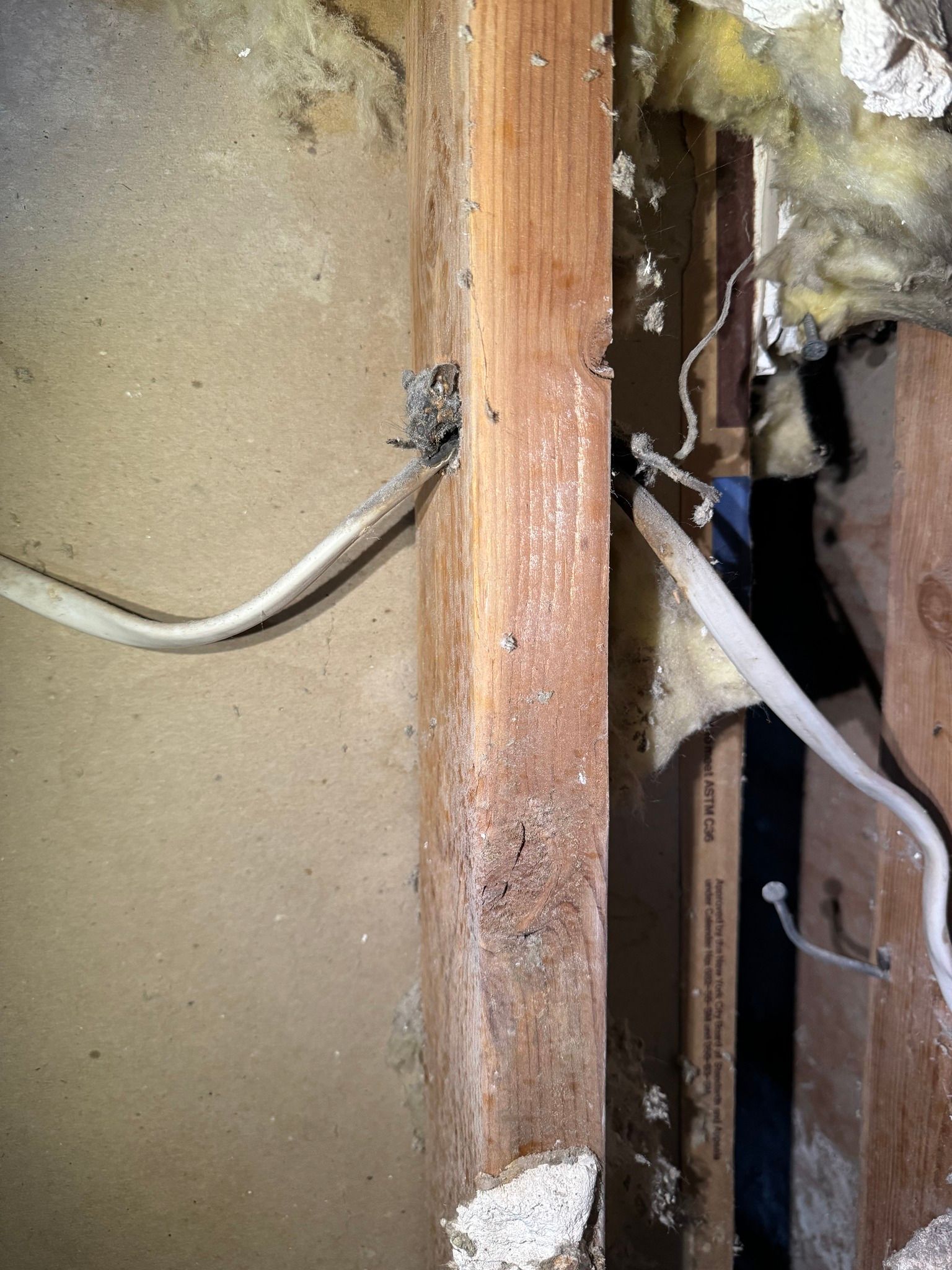 Electrical wiring runs through a hole in a wooden wall stud, with additional wiring and insulation visible nearby.