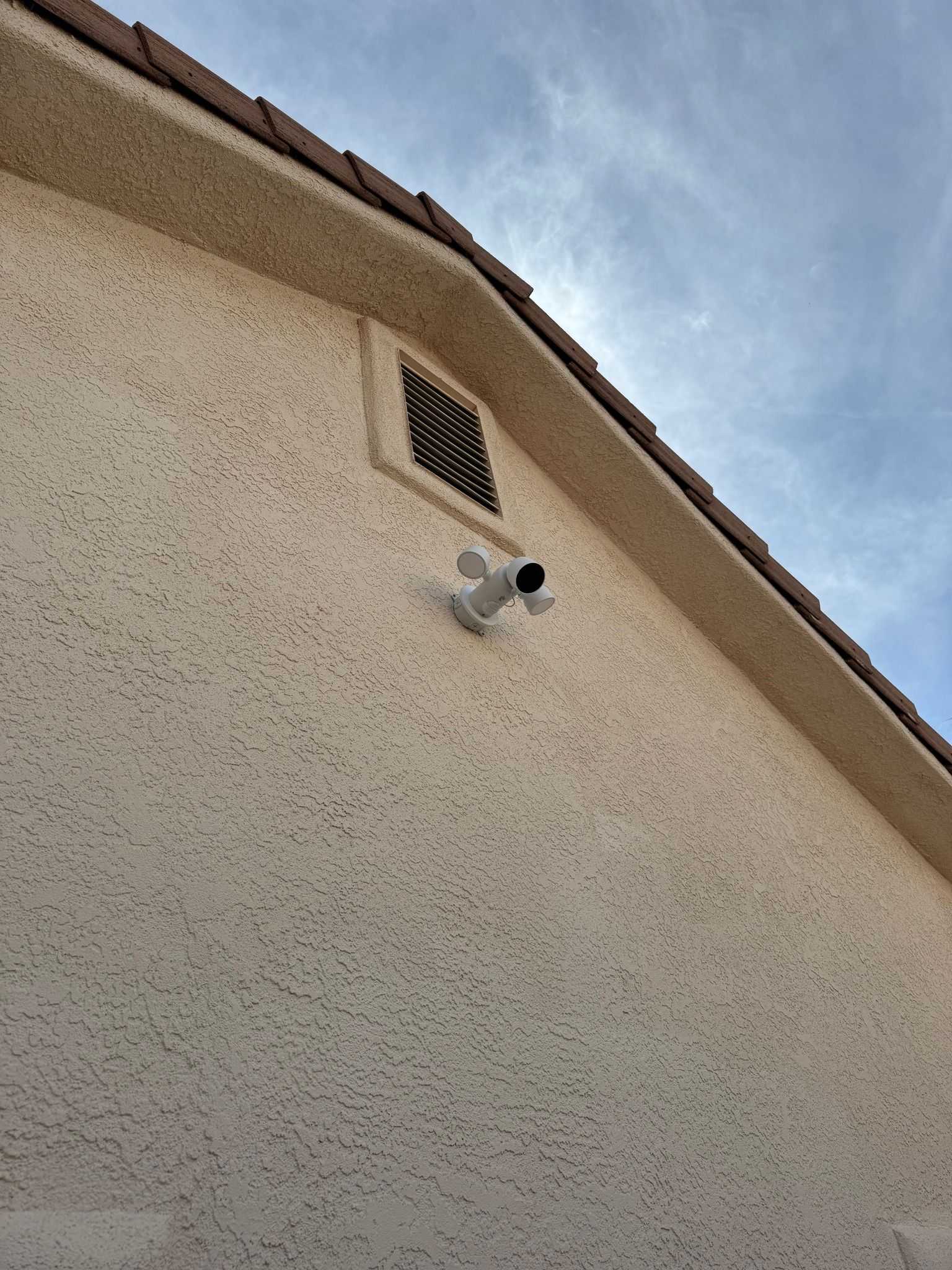 A white security camera with two side-mounted floodlights mounted on a beige, stucco-textured exterior house wall.