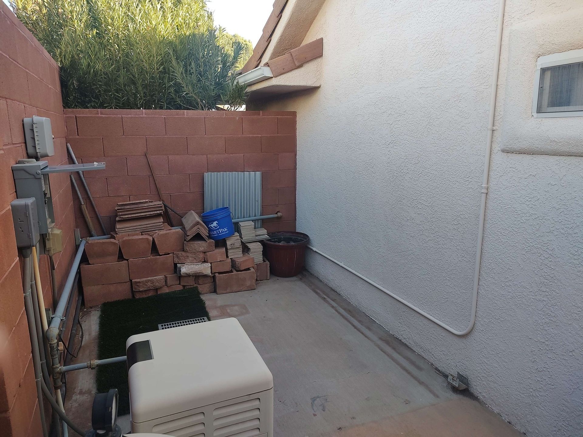A side yard area with a concrete floor, a tan equipment cover, a stack of bricks, and a stucco house wall.