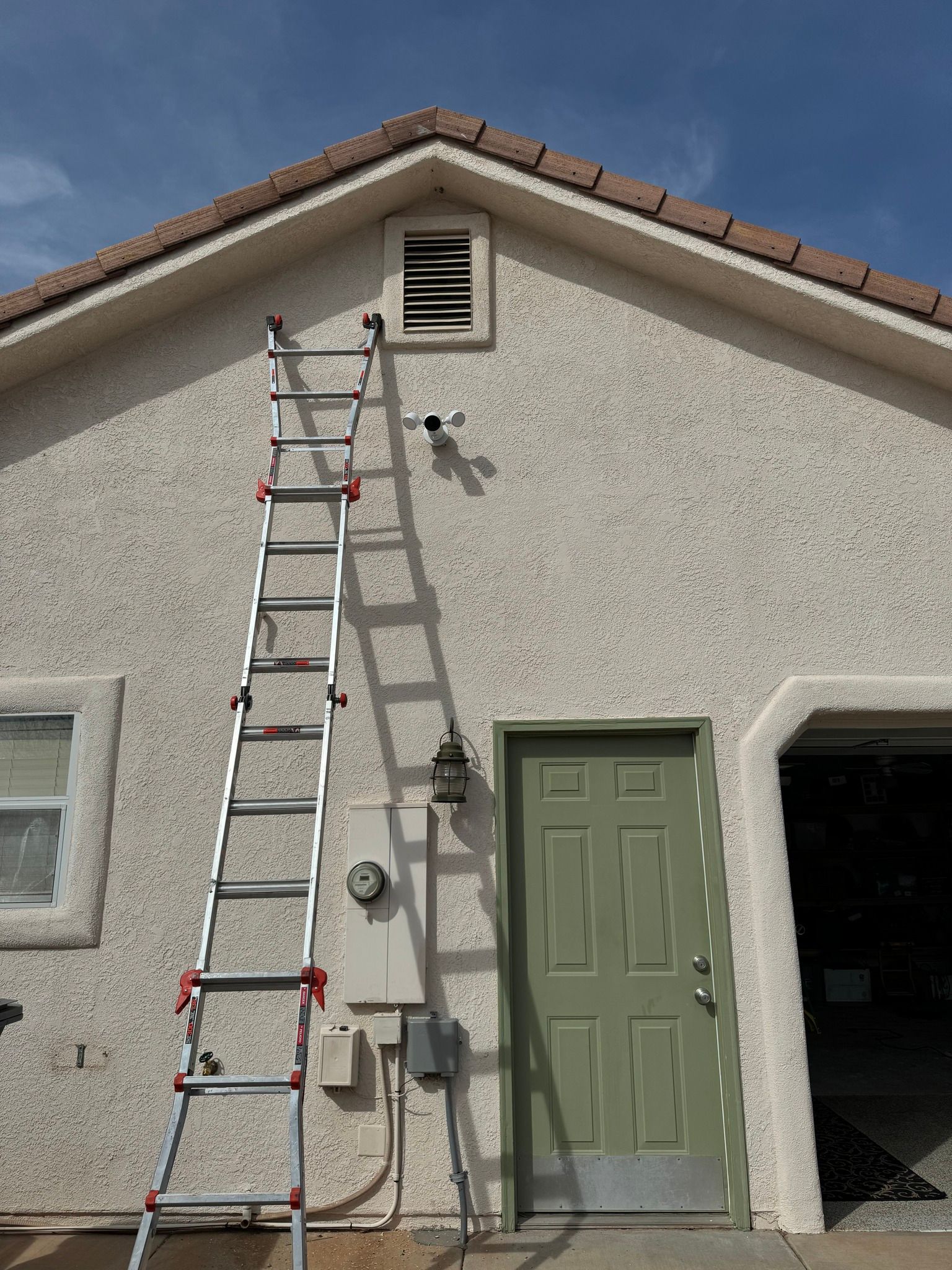 An extendable ladder leaning against the stucco exterior wall of a house near a green door and an attic vent.