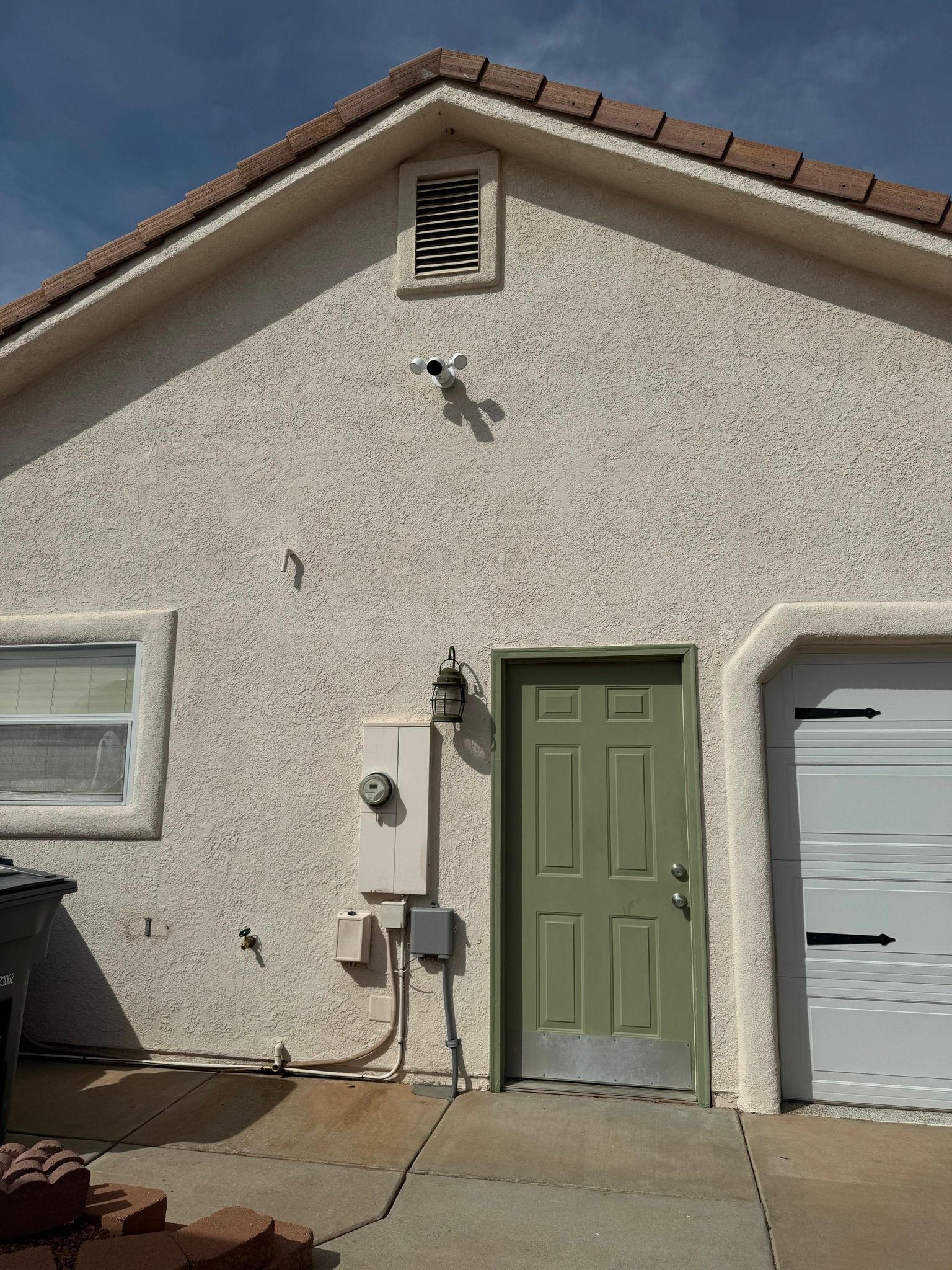 Exterior of a house wall with stucco finish, a green door, a small window, electrical box, and a security camera.
