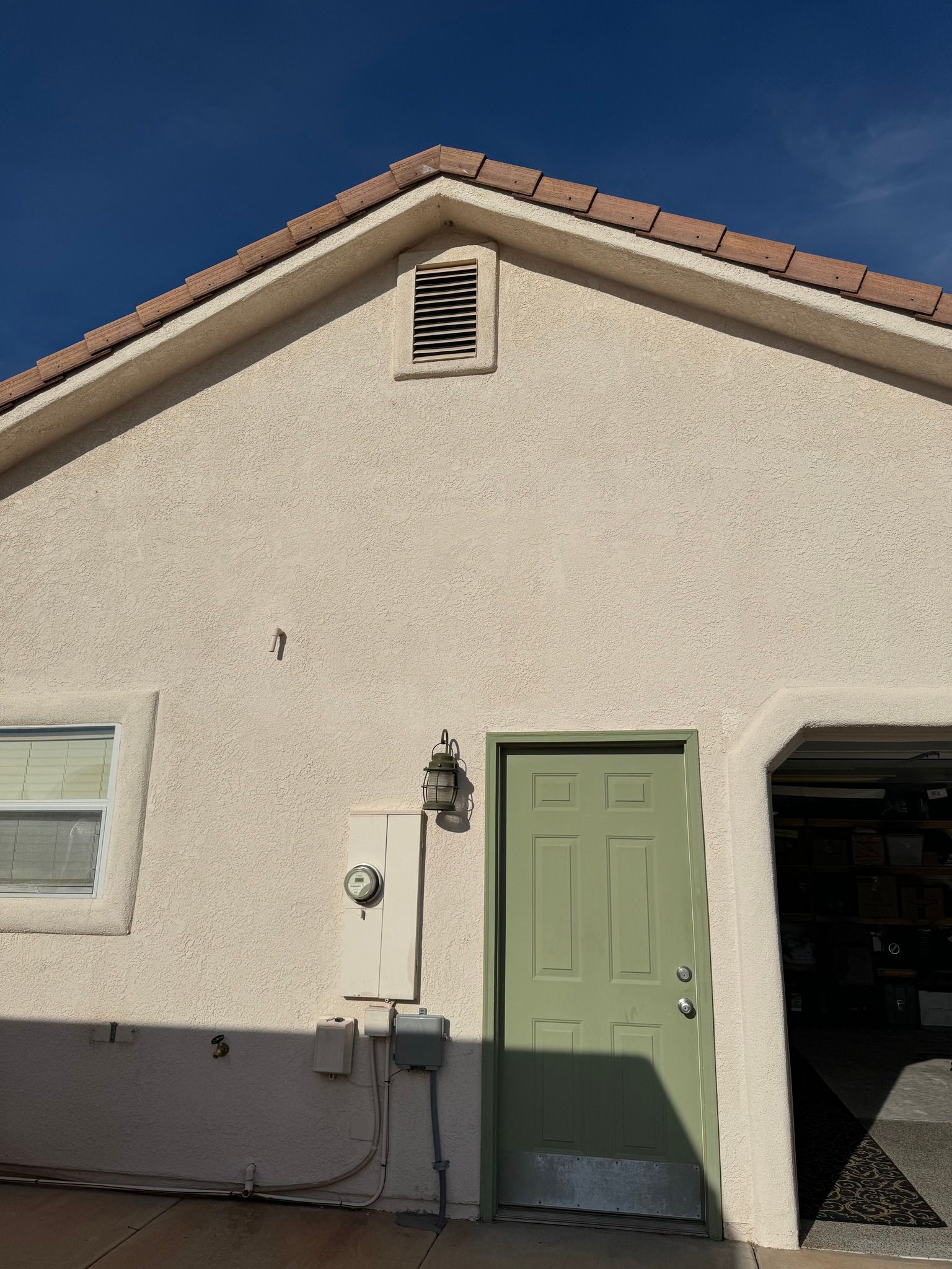 A beige stucco exterior wall with a green door, a small window, an electric meter, and a vent under the roof peak.