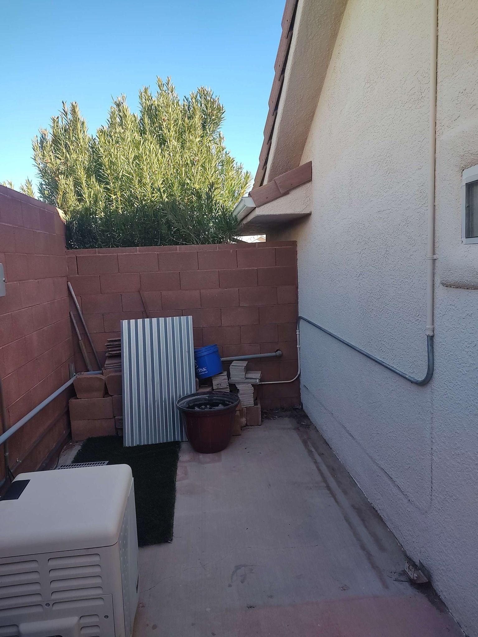 An outdoor side yard with a concrete patio, a beige HVAC unit, a block wall, and some stacked materials near a house.