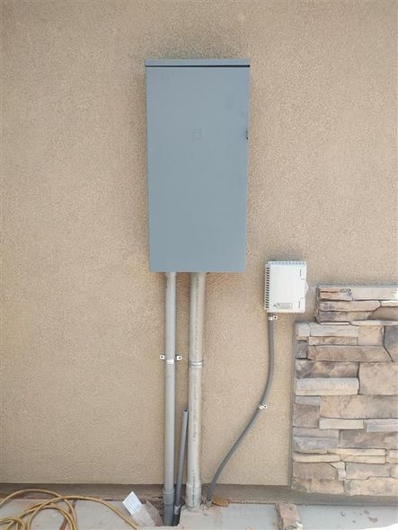 A large grey electrical panel mounted on a beige stucco wall, with a smaller white communication box to its right.