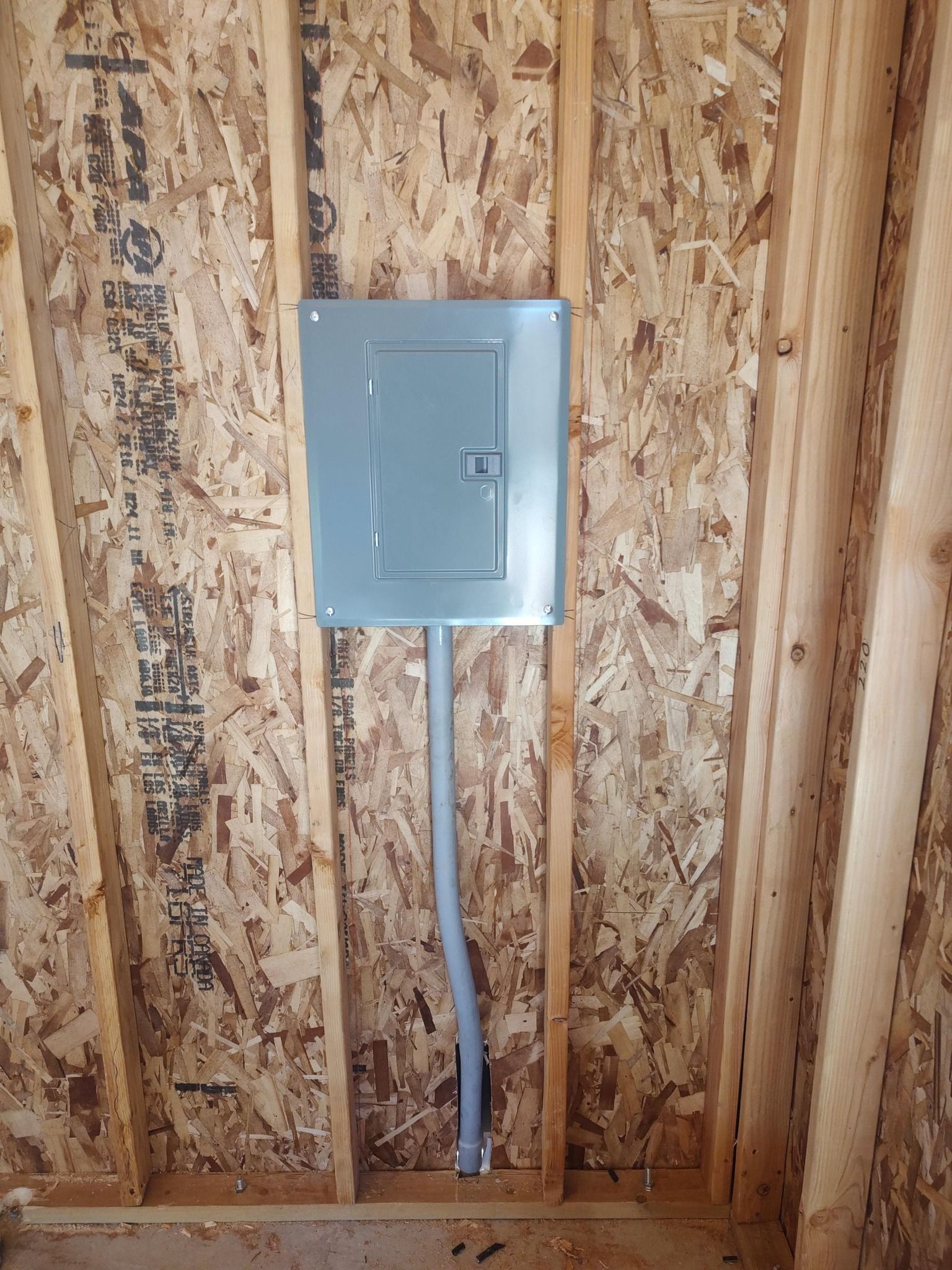 A grey electrical breaker panel mounted between two wooden wall studs with a grey conduit extending downward.