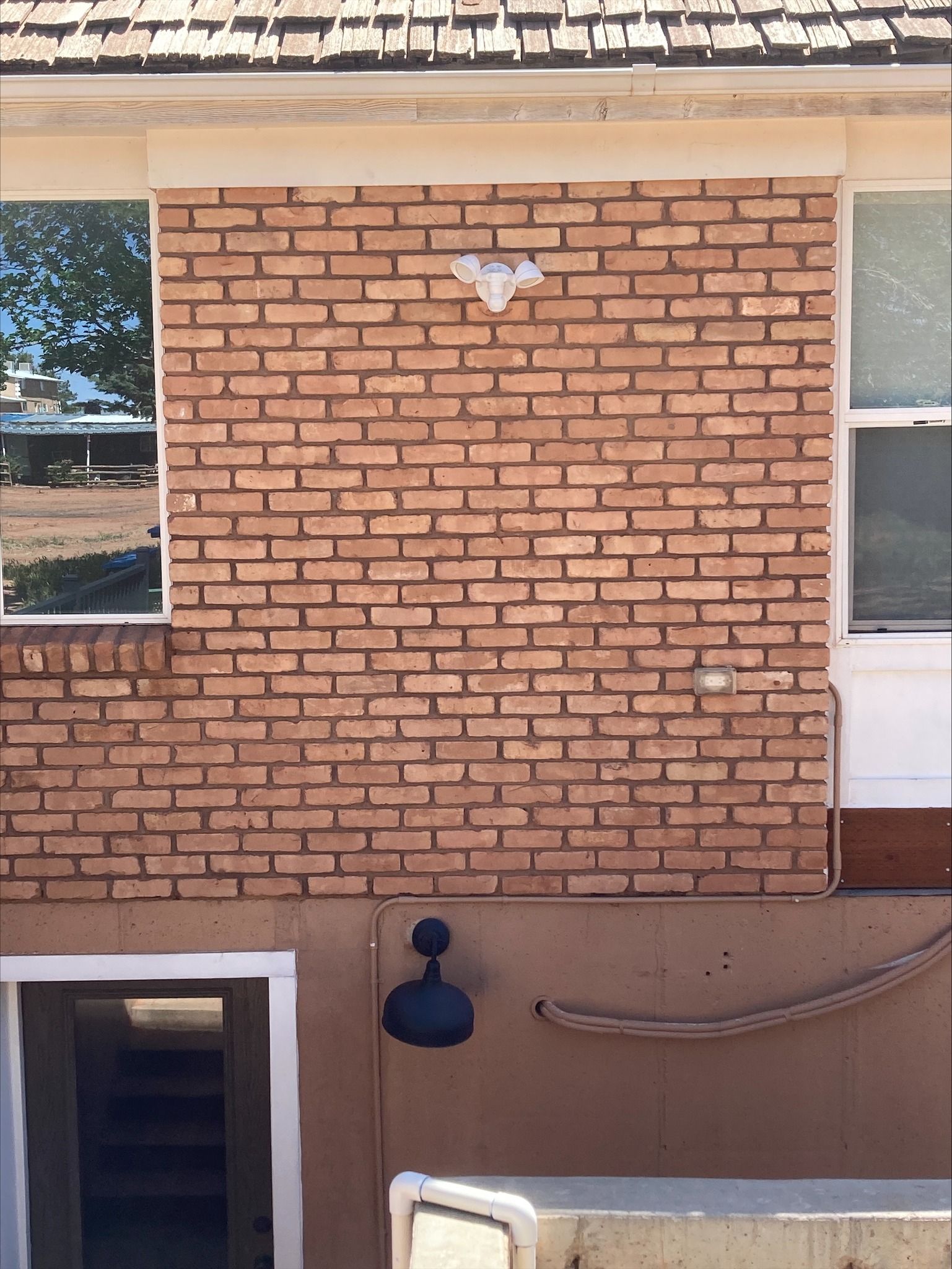 An exterior view of a brown brick wall between two windows, featuring a security light and an outdoor light fixture below.