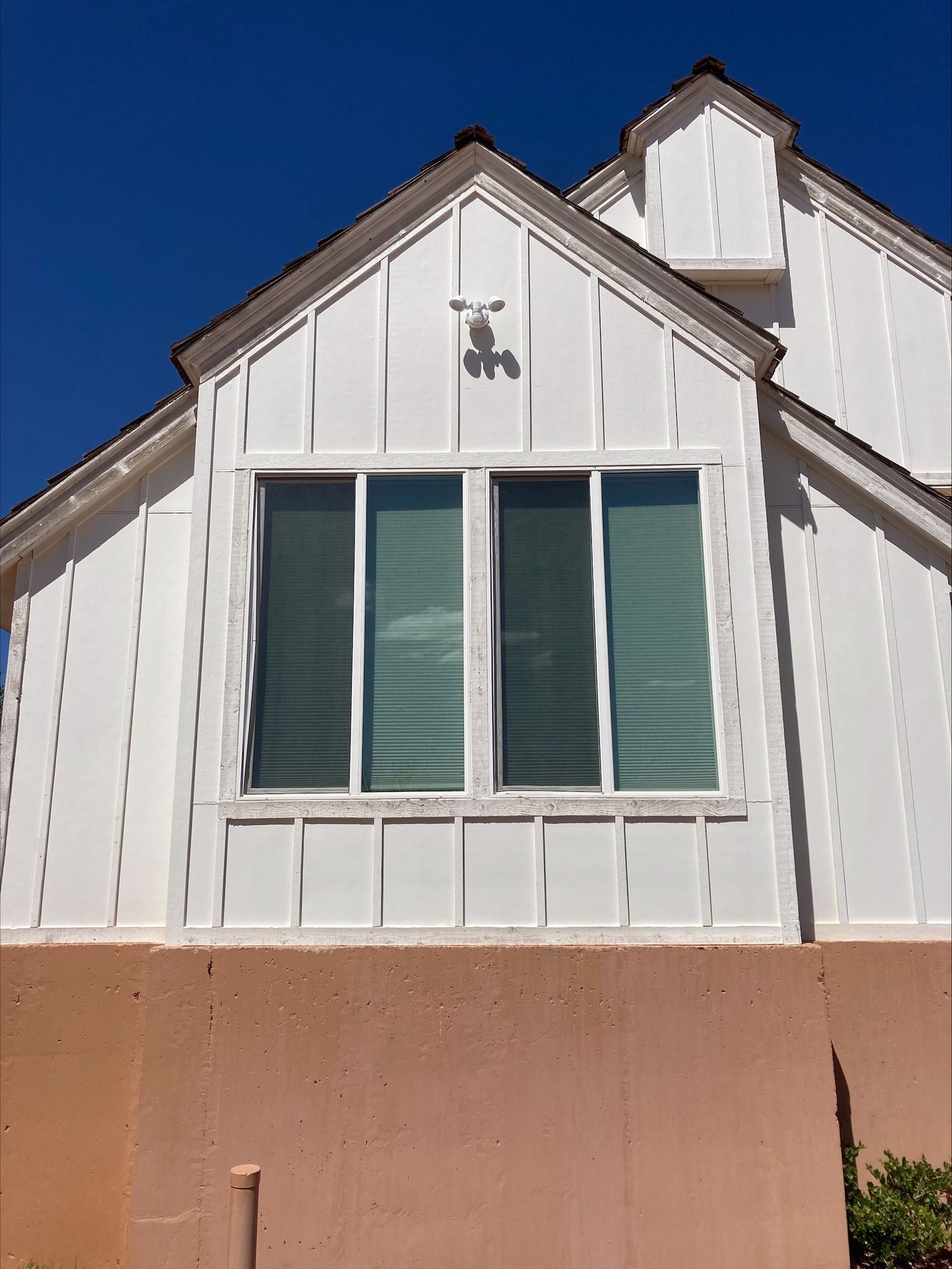 A white, board-and-batten-style house exterior with a large window and a peach-colored foundation under a clear blue sky.