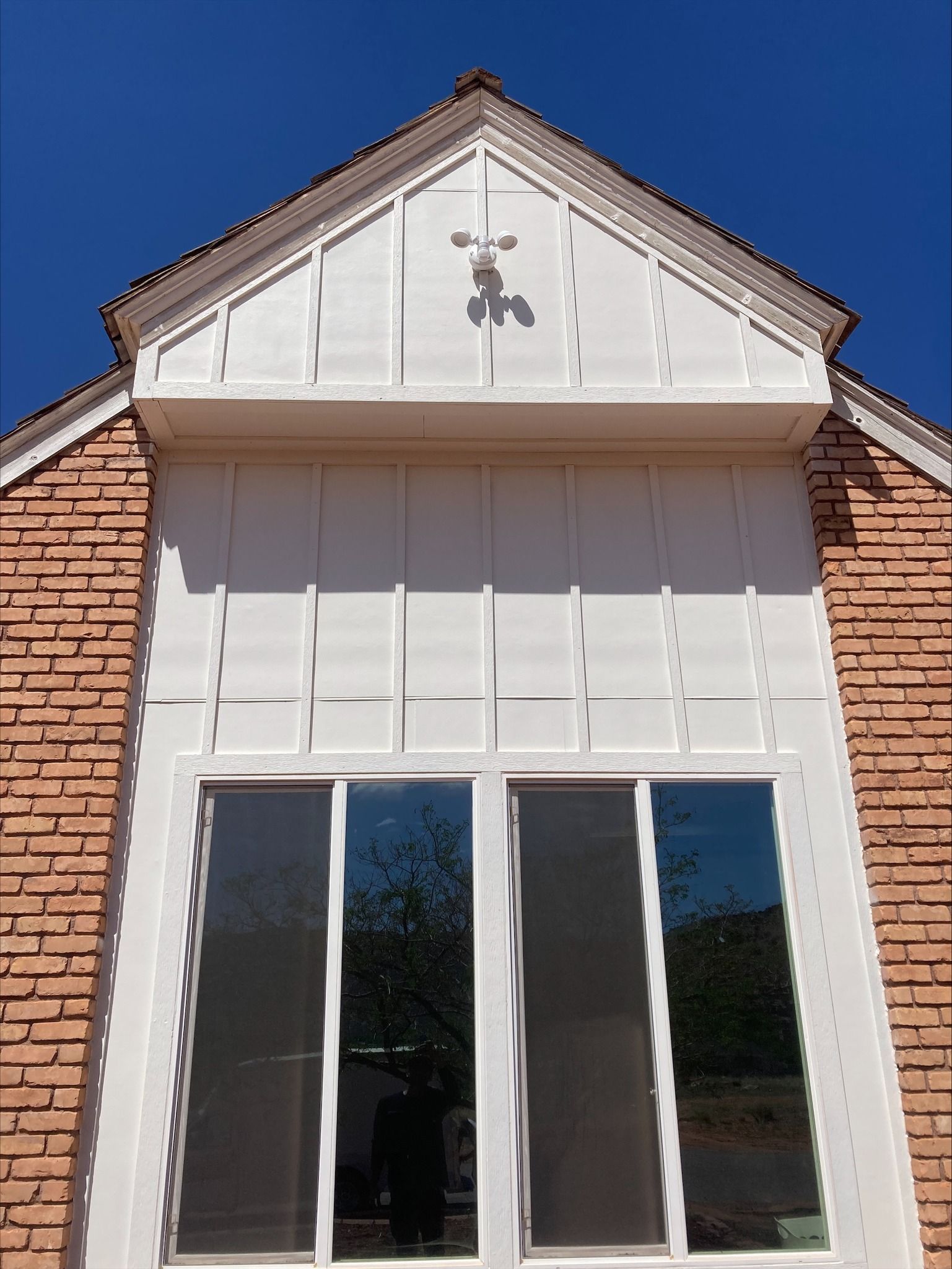 A white, gable-roofed section with vertical siding above a large window, framed by reddish-brown brick walls under blue sky.