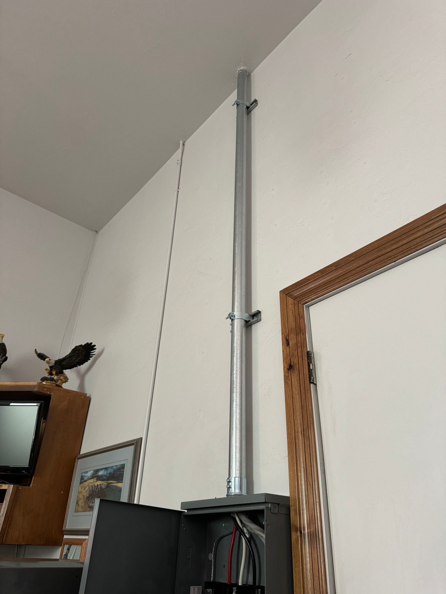 A metal electrical conduit runs vertically up a white wall to the ceiling above an open electrical panel.