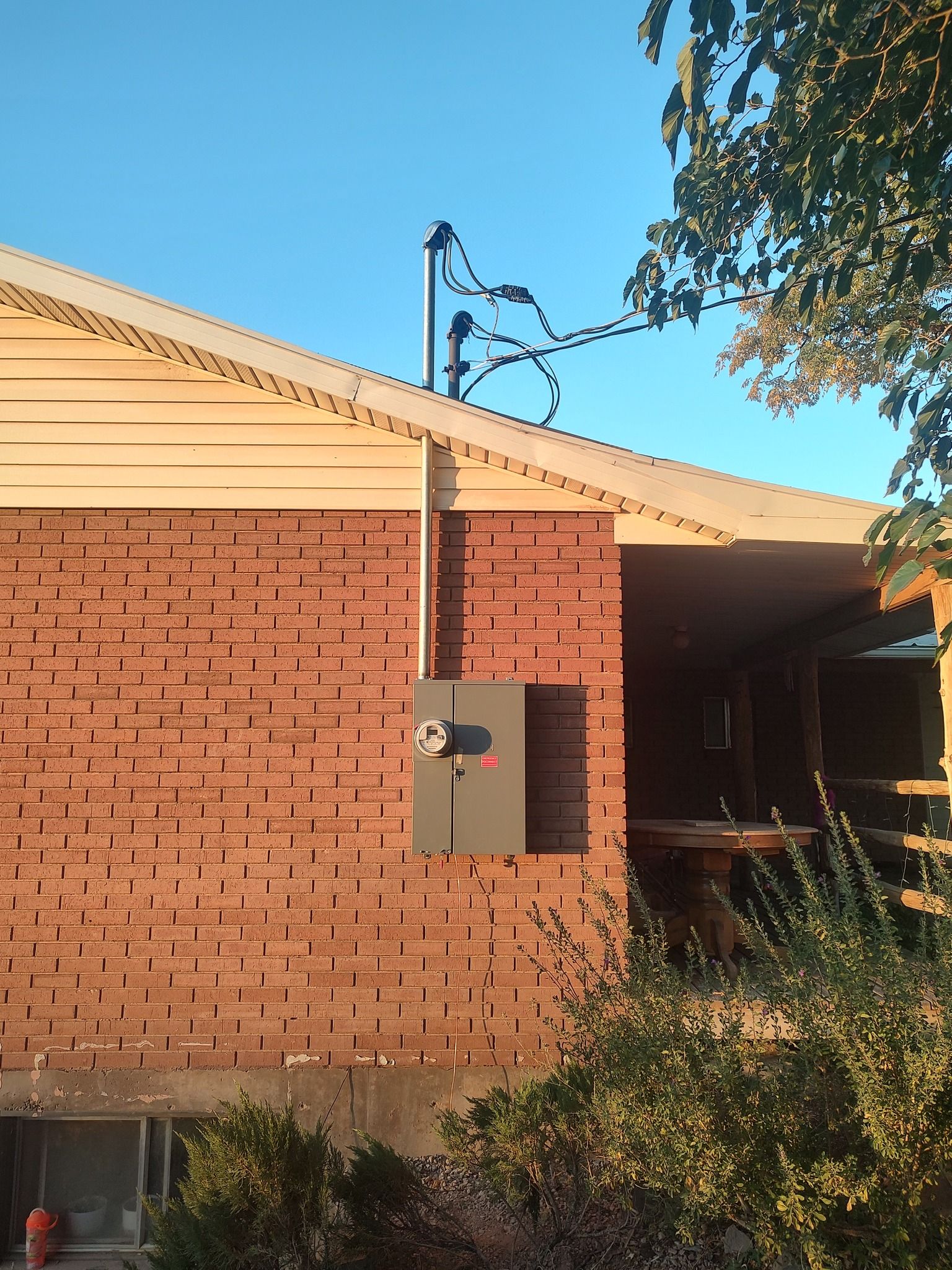 A brick house exterior showing an electrical service mast connected to a gray meter base box mounted on the wall.