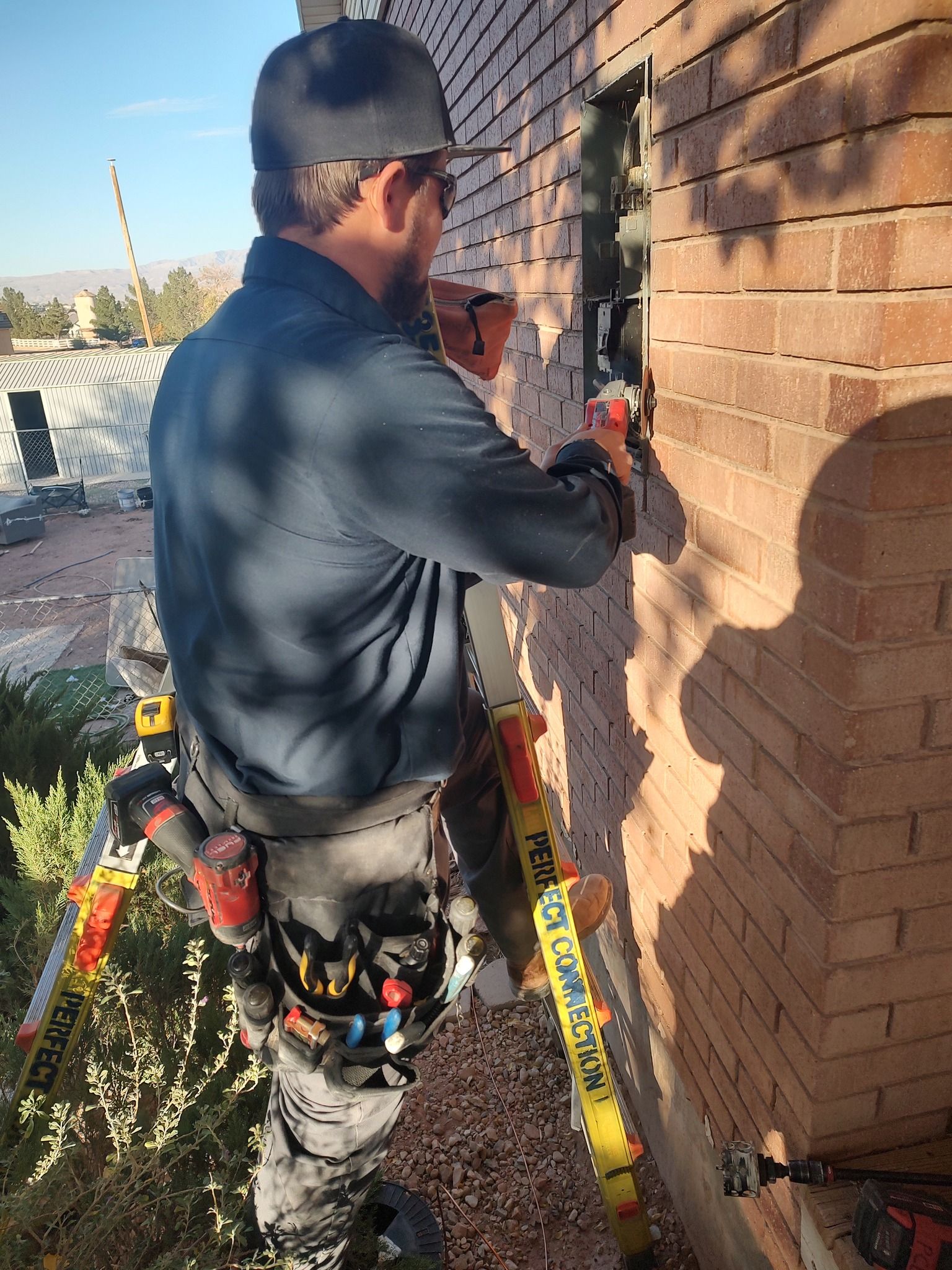 A worker in a dark shirt and tool belt stands on a ladder, installing electrical components into a brick wall.