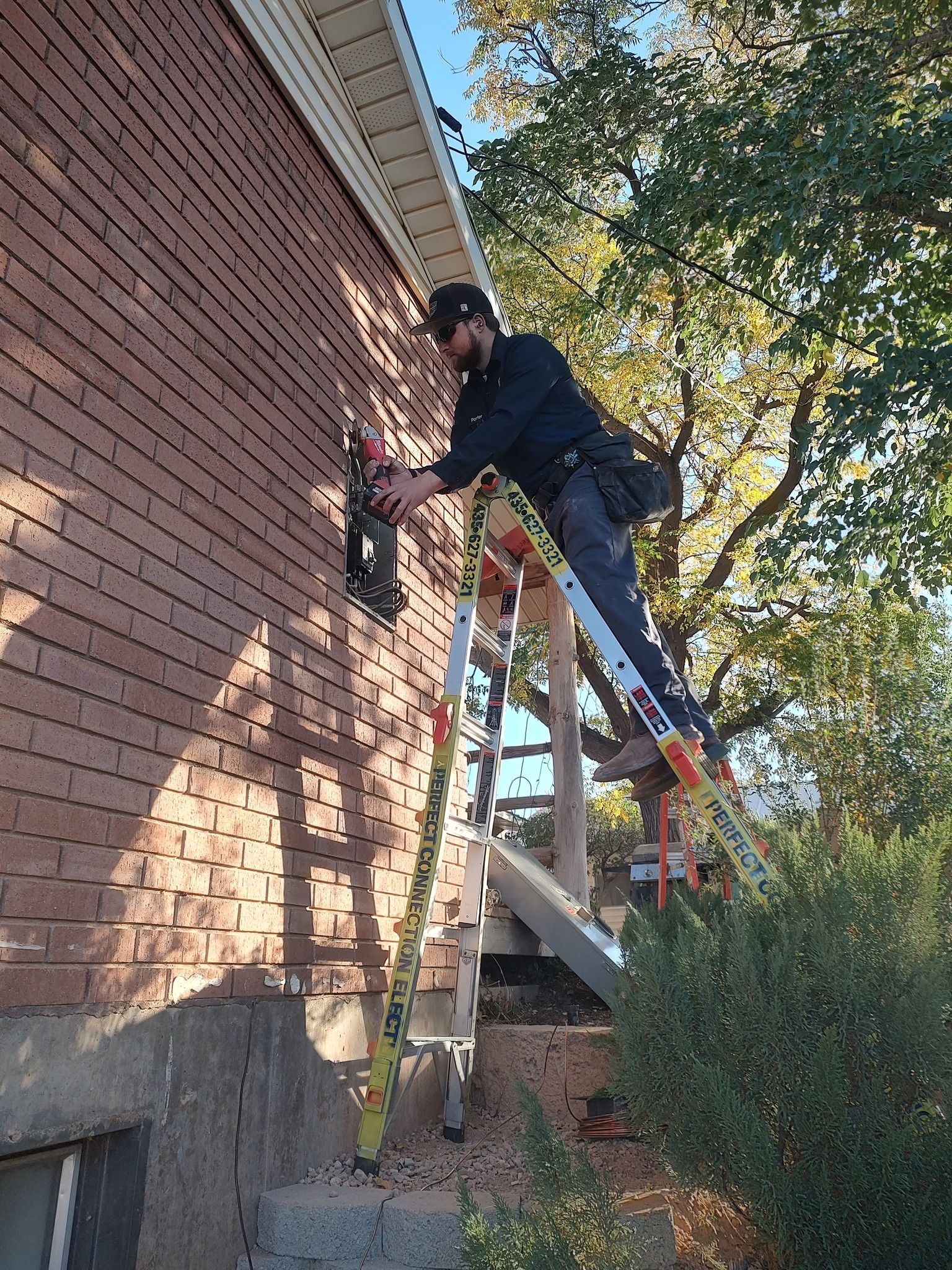 A person in dark clothing stands on a tall ladder, working on a brick wall exterior of a house during the day.