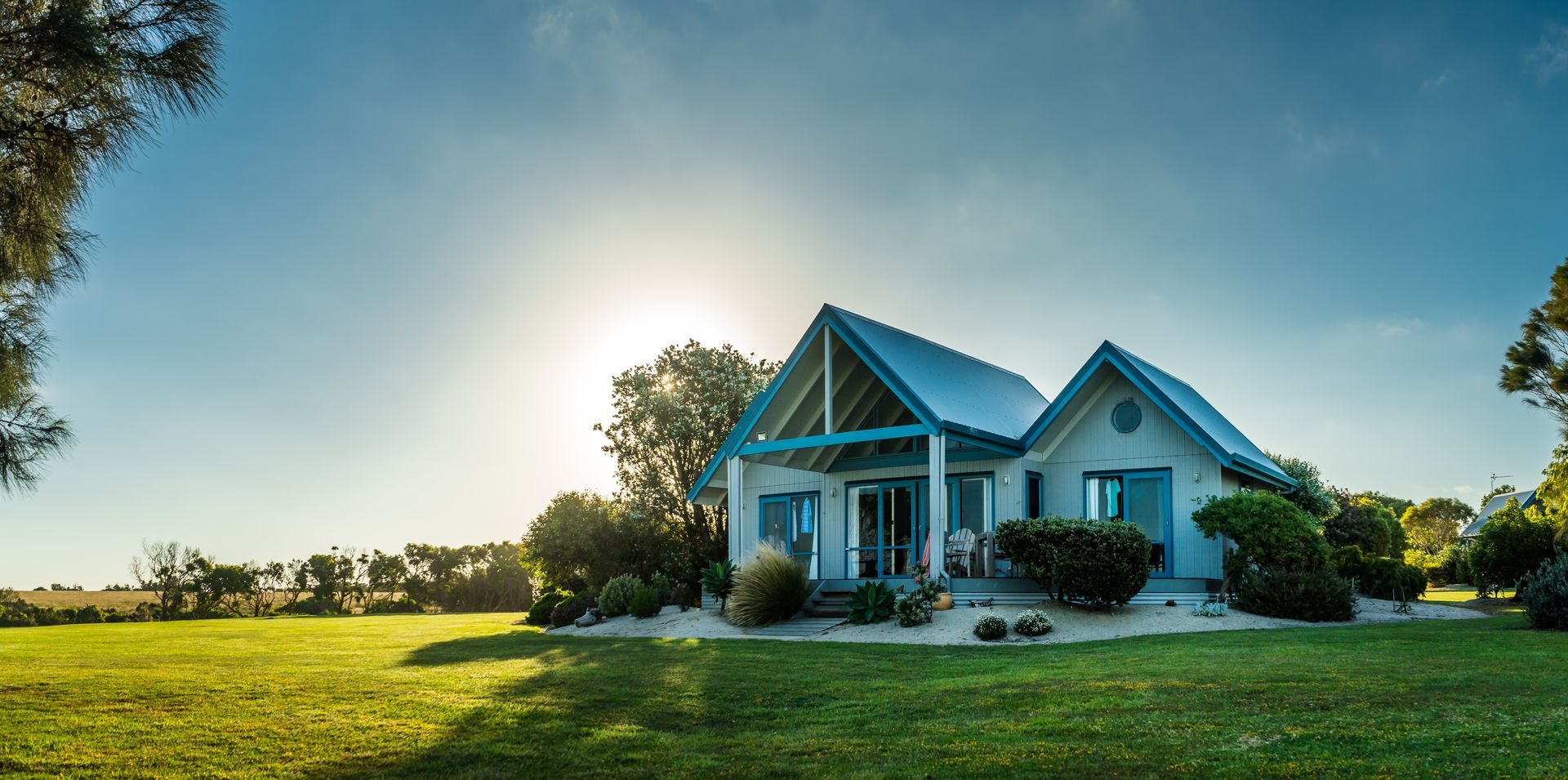 Une maison bleu clair se dresse au milieu d'un champ verdoyant sous un ciel dégagé, baigné de soleil.