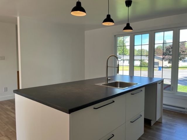 Îlot de cuisine moderne avec évier et suspensions lumineuses. Placards blancs, plan de travail foncé et grande fenêtre.