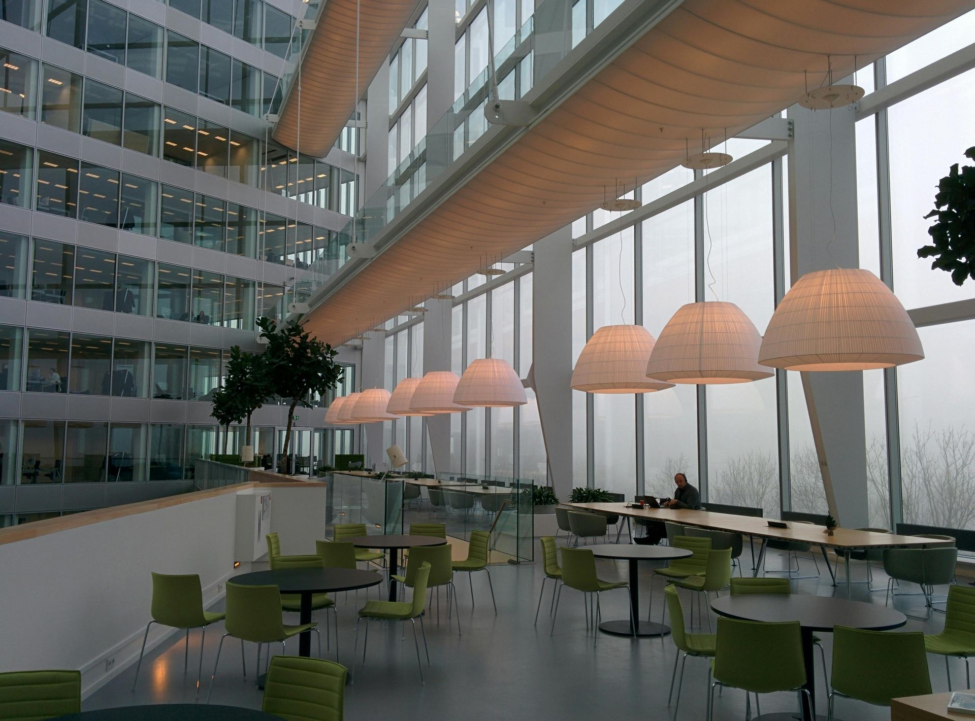 Une cafétéria de bureau avec des tables rondes, des chaises vertes et de grandes fenêtres.