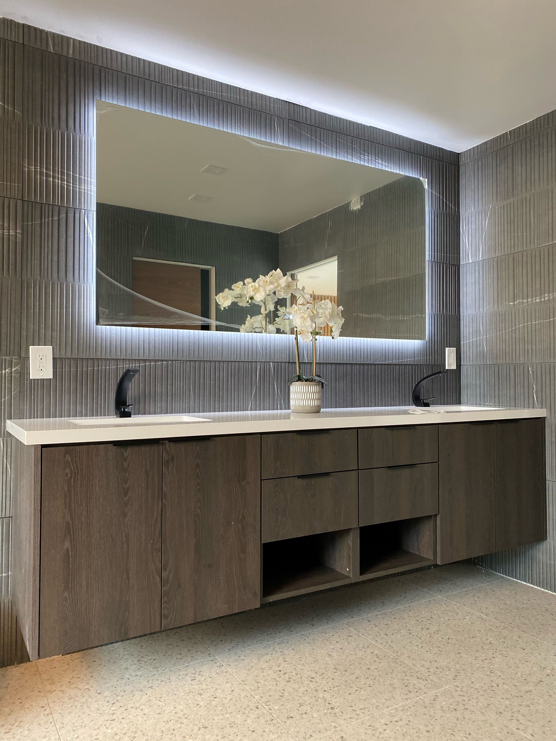 Modern bathroom with a large lit mirror, double vanity, and gray textured tile.