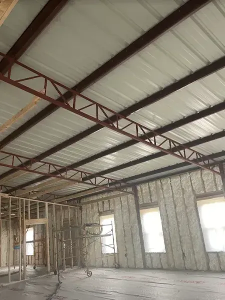 Interior of a building under construction featuring exposed steel roof trusses, metal ceiling panels, and spray insulation.