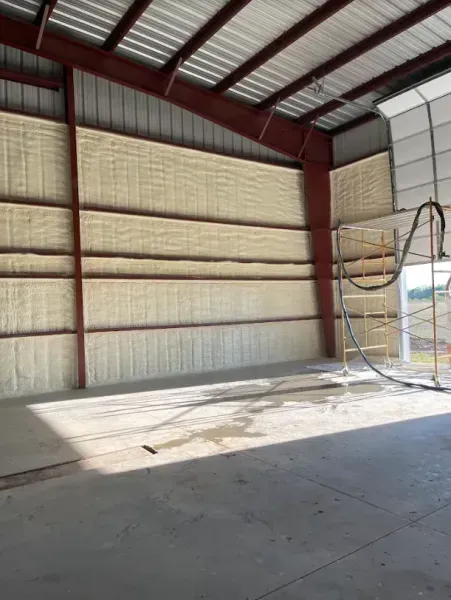 A large metal workshop interior featuring walls recently insulated with thick, tan spray foam, with scaffolding visible.