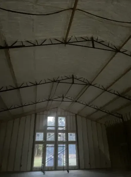 Interior of a large building with high ceilings and walls covered in white spray foam insulation and a large window.