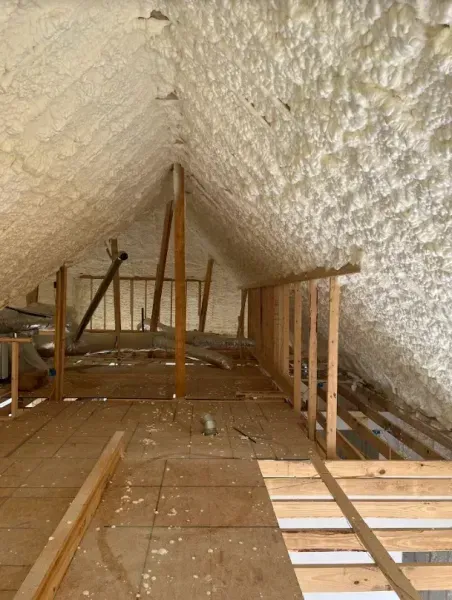 An attic space with light-colored spray foam insulation on the slanted roof, wooden flooring, and partial wall framing.