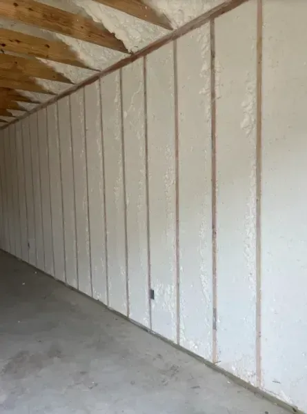 An interior view of a garage wall framed with vertical wooden studs and filled with white spray foam insulation.