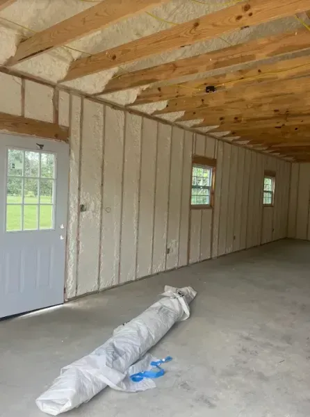 An interior view of a room under construction with spray foam insulation on the walls, wooden ceiling beams, and a door.