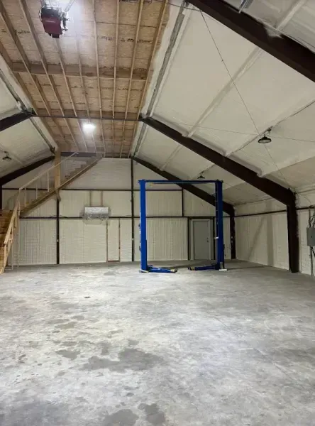An empty warehouse or garage space with a blue two-post car lift, a wooden staircase, and exposed ceiling beams.