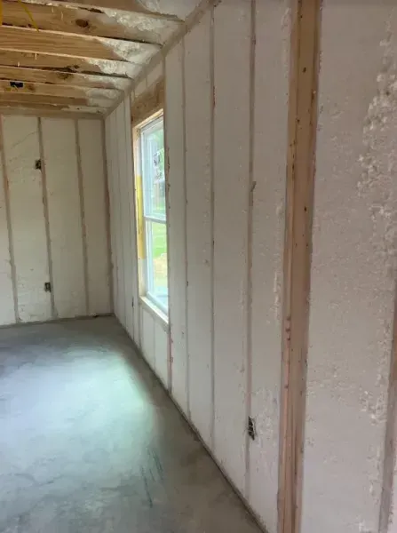 Interior view of an unfinished room with wooden wall studs, spray foam insulation, a single window, and concrete floors.