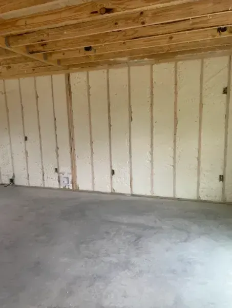 Interior view of an unfinished room with concrete floors, exposed wooden ceiling joists, and spray-foam insulated walls.