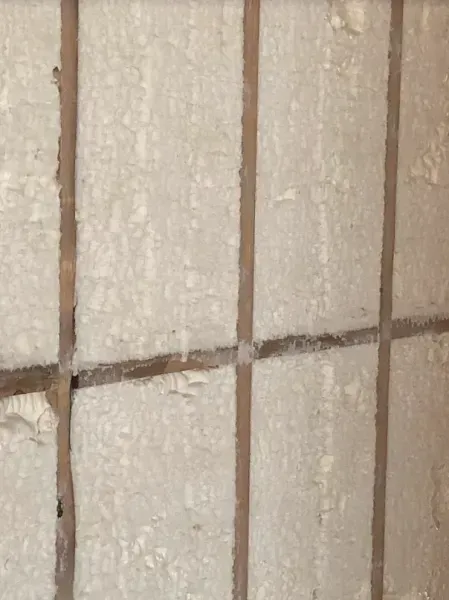 Close-up of a wall featuring a grid of thin, brown wooden strips framing panels of rough, light-colored insulation.