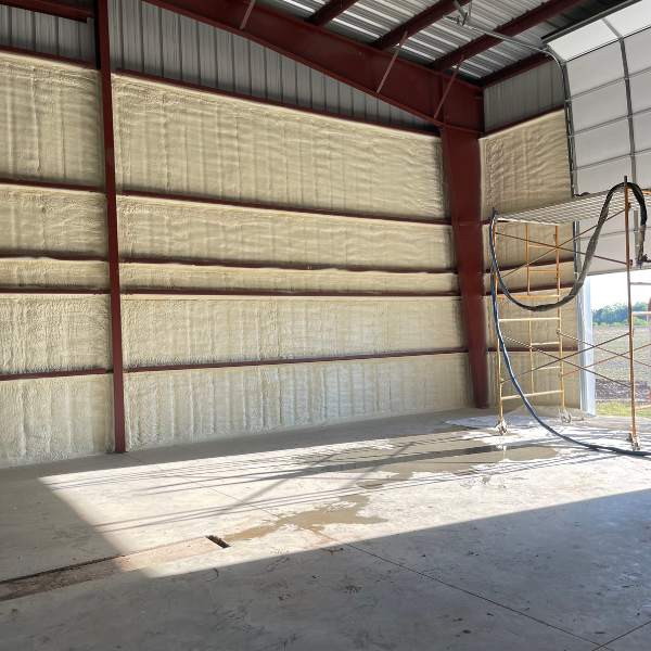 Interior view of a large metal building with walls insulated by sprayed yellow foam, featuring scaffolding to the right.
