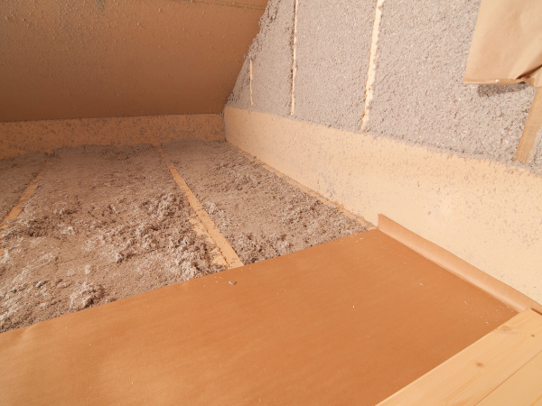 Attic space with light brown wooden floorboards and gray loose-fill insulation between wooden ceiling joists.