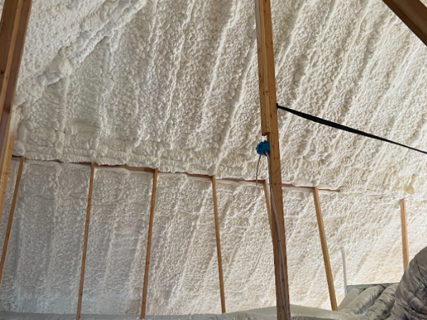 Attic rafters and walls covered in white spray foam insulation, with wooden support beams and an exposed blue electrical box.