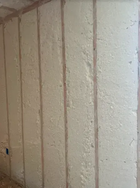 A wall featuring vertical wooden studs with gaps filled with beige spray foam insulation.
