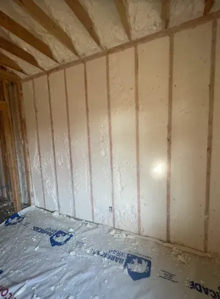 An interior room under construction featuring wood wall studs insulated with white spray foam and floor sheeting.