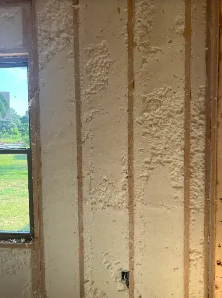 Interior wall framing with spray foam insulation applied between the wooden studs, next to a window.