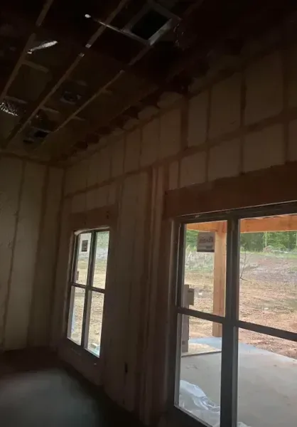 Interior view of a residential room under construction, showing wooden wall framing, open ceiling joists, and two windows.