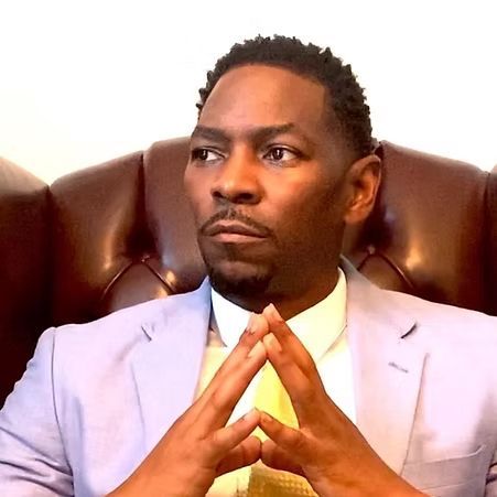Man in a light blue suit sitting in a brown leather chair, hands steepled, looking to the side.