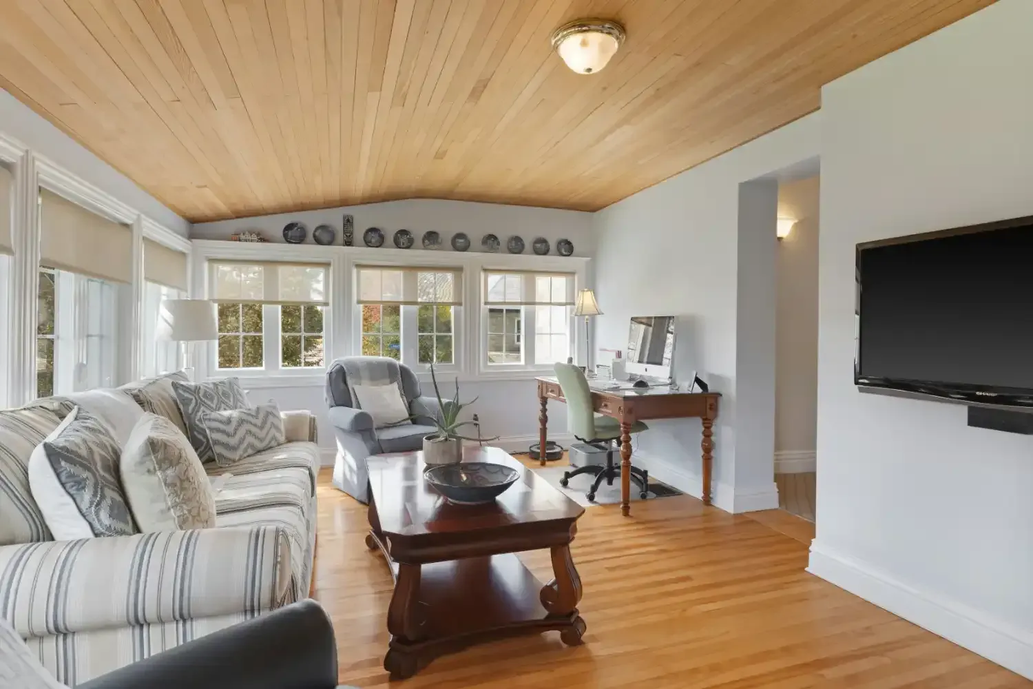 Bright living room with striped sofas, wooden ceiling, wall-mounted TV, and large windows.