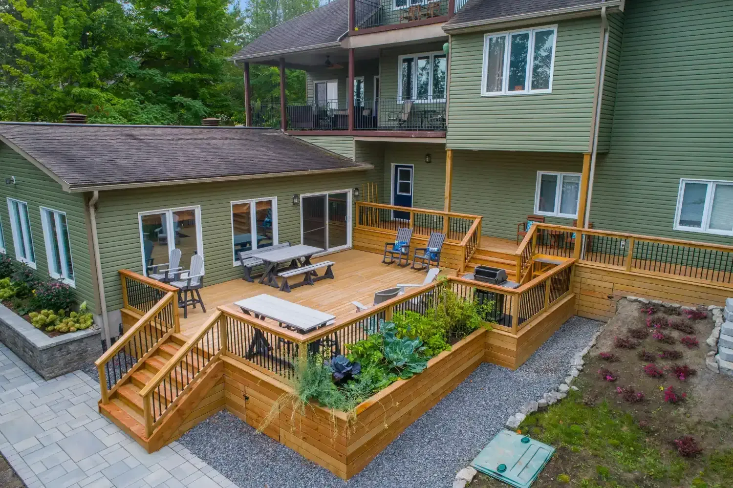 Large wooden backyard deck with stairs, benches, table, and planters beside a green house