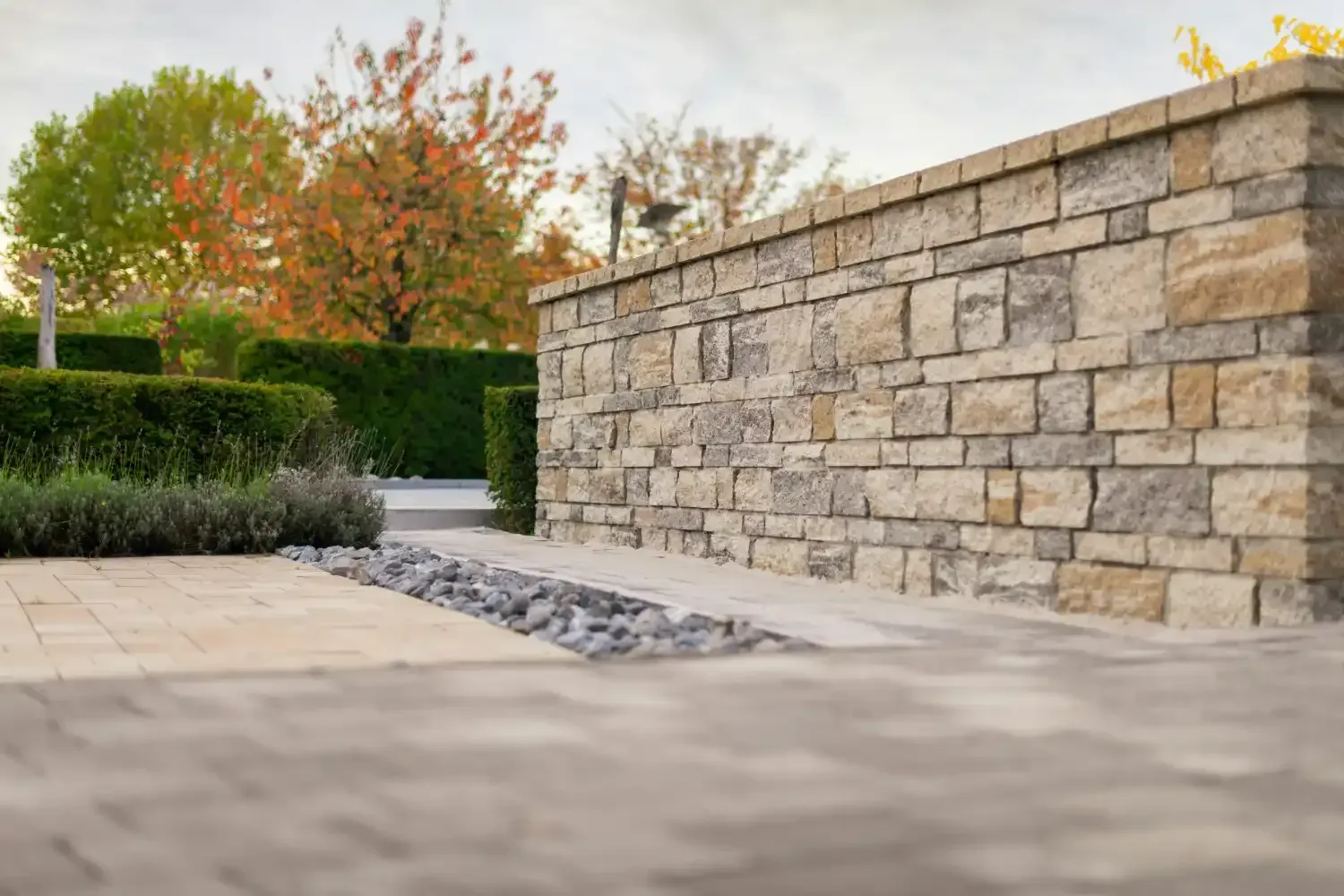 Stone retaining wall beside a paved walkway, with trimmed hedges and autumn trees in the background.