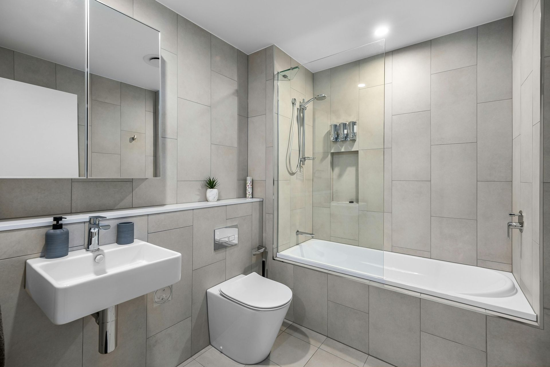 Modern bathroom with white vanity, toilet, mirror, and a bathtub-shower combo in light gray tones