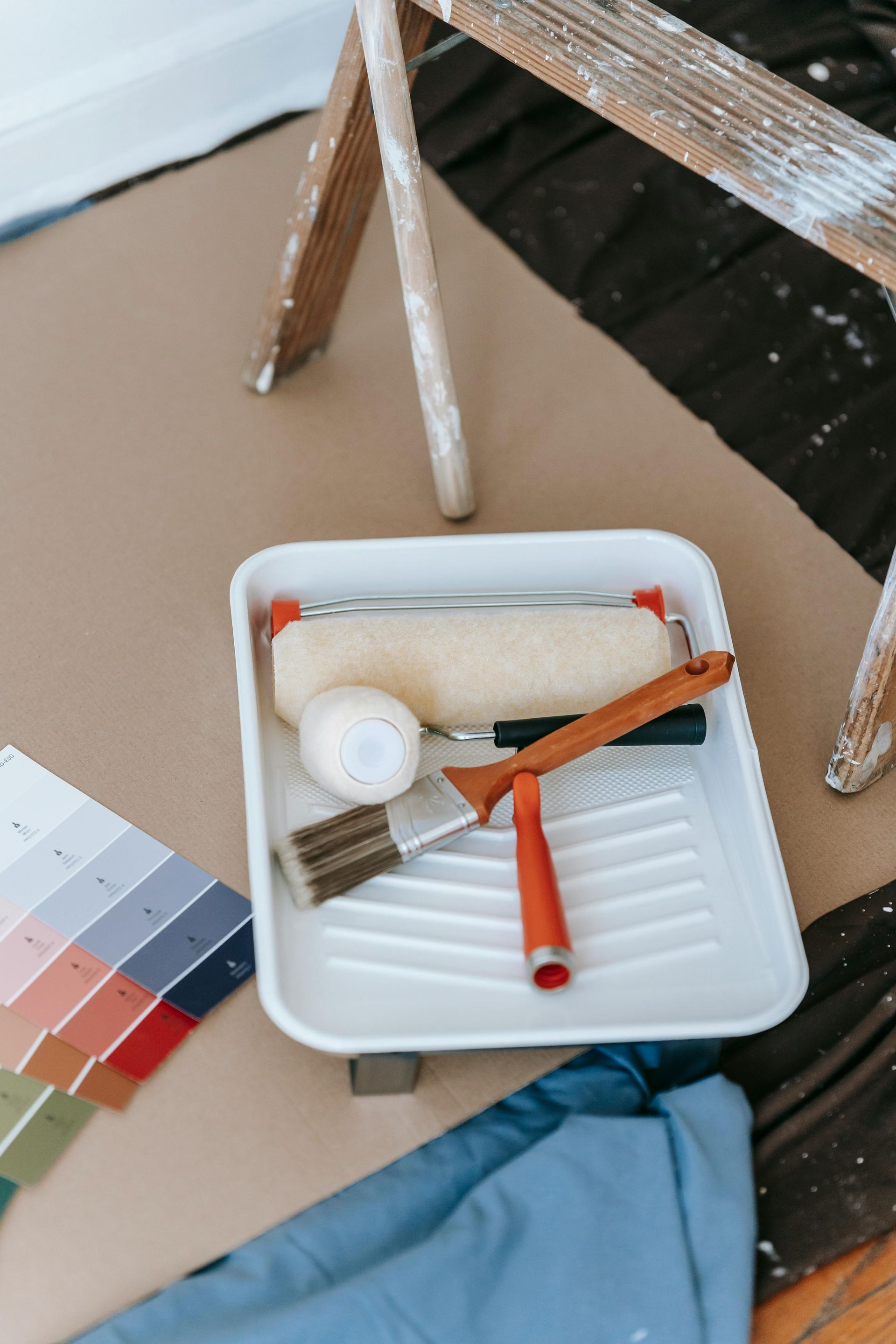 Paint supplies in a white tray on a table, with color swatches, a ladder, and fabric nearby.