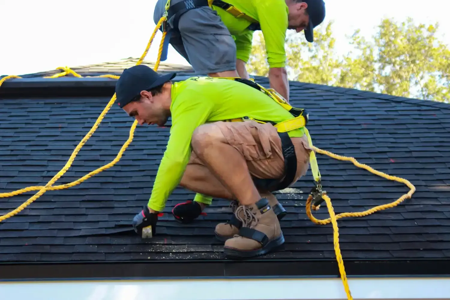 Two roofers in bright shirts securing yellow safety ropes on a shingled roof