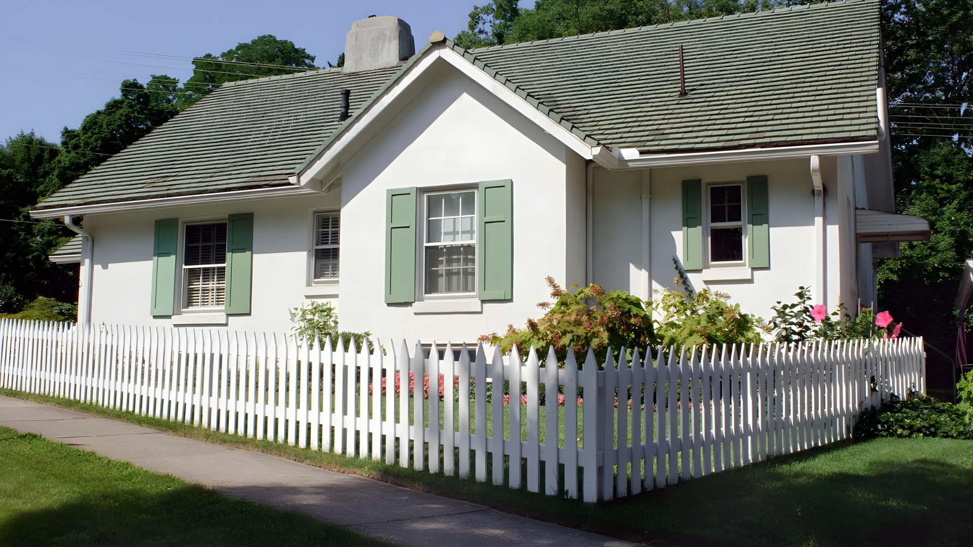 White cottage with green shutters and a picket fence in a sunny garden