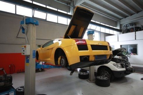 Yellow sports car lifted on a hydraulic hoist in an auto repair shop, with a second car below