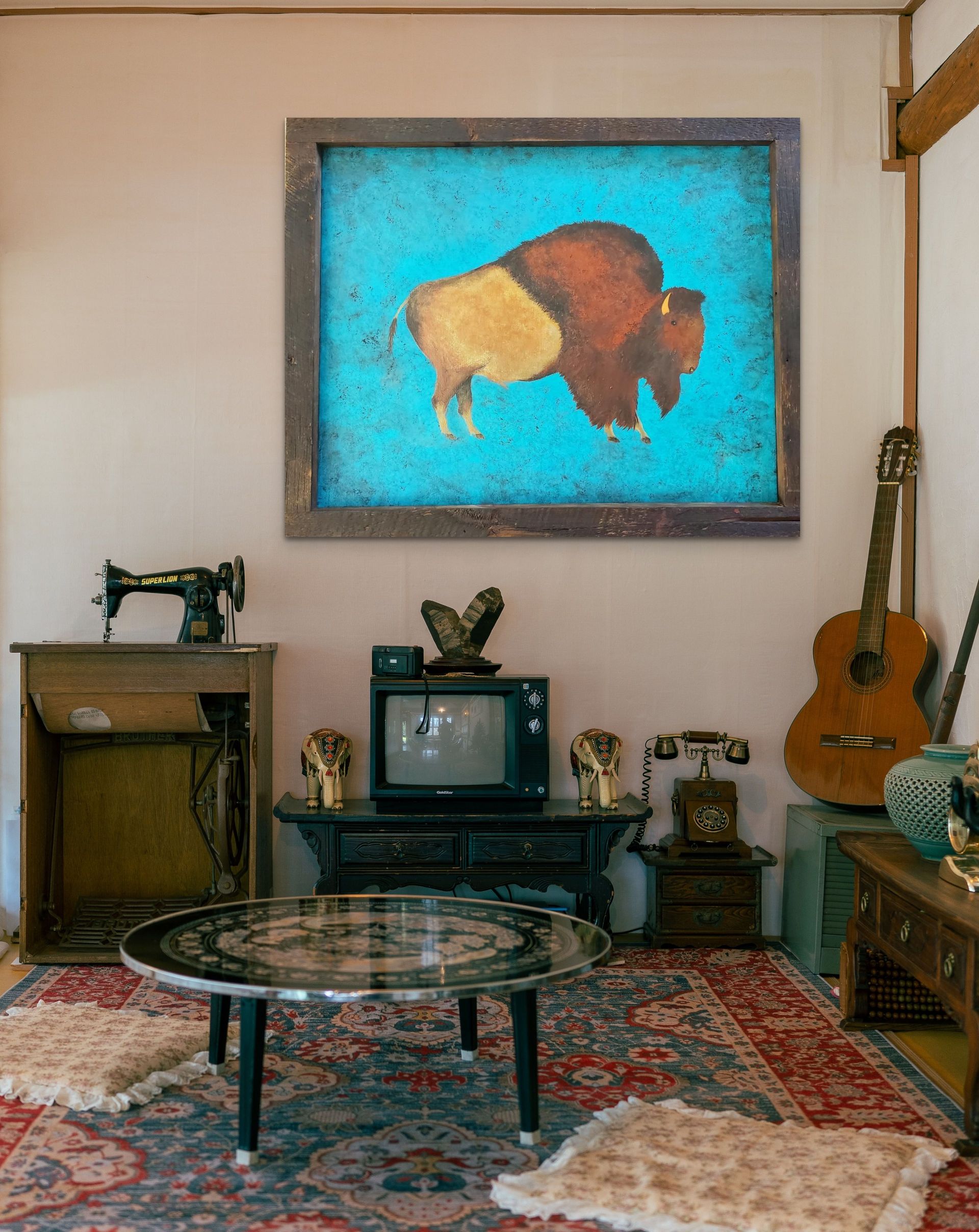 A vintage-style room with a bison painting, sewing machine, old TV, rotary phone, and acoustic guitar on a rug.