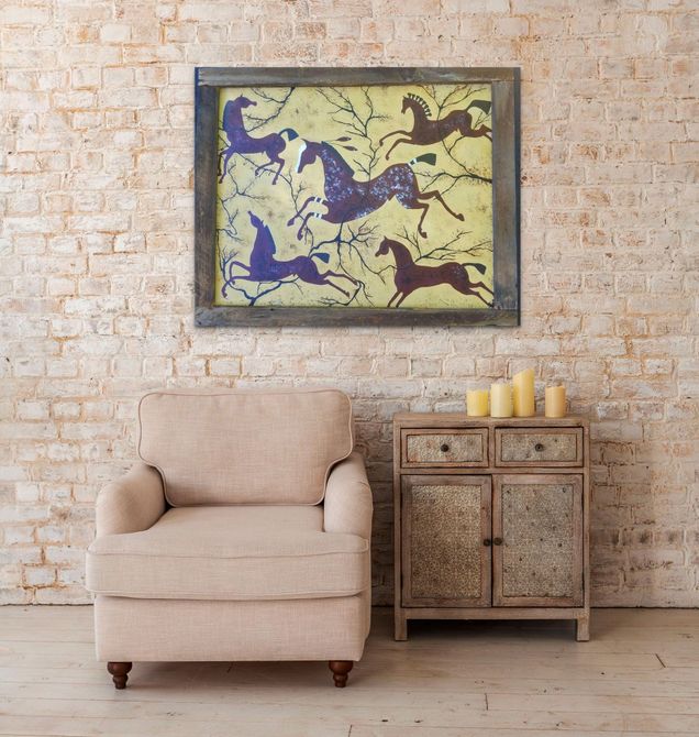 An armchair and a small wooden cabinet with candles sit against a brick wall beneath a framed painting of horses.